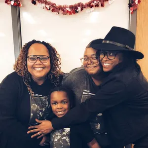 Bernice Hardman poses with three family members, all wearing D'Angelo Black Messiah shirts, in front of a whiteboard decorated with red Valentine's Day garland.