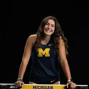 Clare McNamara smiles while leaning on a hurdle branded with "Michigan," photographed against a black background.A young woman smiles while leaning on a hurdle branded with "Michigan," photographed against a black background. She wears a navy blue University of Michigan athletic tank top with the block M logo in maize and gold.