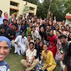 A large group of students and faculty smile for a selfie outdoors on a college campus, with a yellow building and trees visible in the background. The diverse group includes participants wearing lanyards with "Madras Christian College" visible on some, alongside others wearing University of Michigan Social Work apparel. Several people flash peace signs, and the mood is celebratory and warm.