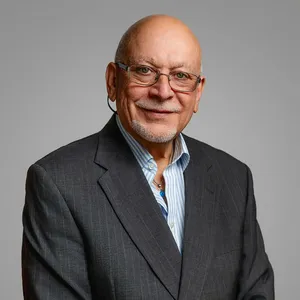 A professional headshot of Osvaldo Rivera wearing glasses, a dark gray pinstripe blazer, and an open-collar blue striped shirt. He has a warm smile, a short white goatee, and is photographed against a neutral gray background.