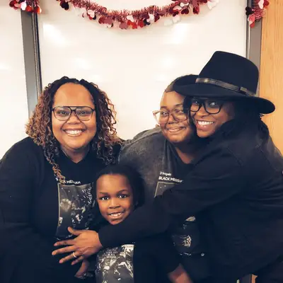 Bernice Hardman poses with three family members, all wearing D'Angelo Black Messiah shirts, in front of a whiteboard decorated with red Valentine's Day garland.