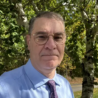 A headshot of Donald Michael wearing round gold-framed glasses, a light blue dress shirt, and a purple patterned tie, photographed outdoors in front of birch trees with green and yellow autumn foliage.