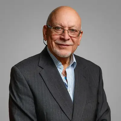 A professional headshot of Osvaldo Rivera wearing glasses, a dark gray pinstripe blazer, and an open-collar blue striped shirt. He has a warm smile, a short white goatee, and is photographed against a neutral gray background.