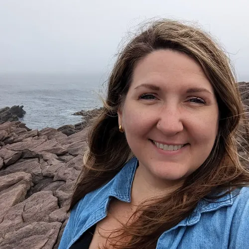 Chelsea Mudalagi smiling outdoors on a rocky coast, wearing a denim shirt.