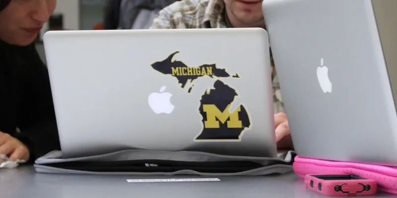 Three students discussing something on a laptop that has a U-M sticker.