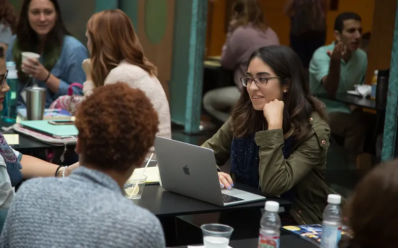 A student at a table in a busy room on a computer listening to someone.