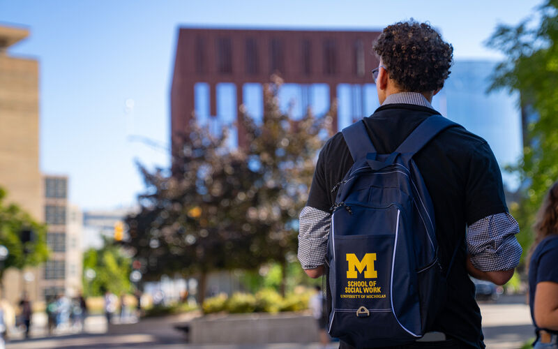 A student seen from behind wearing a navy University of Michigan School of Social Work backpack, looking toward a brick building on a sunny campus day.
