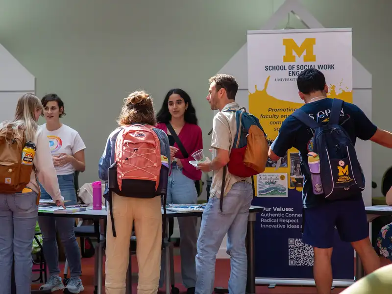 Students carrying backpacks gather around a table at the School of Social Work's student org resource fair, talking with staff and browsing informational materials, with a School of Social Work banner in the background.