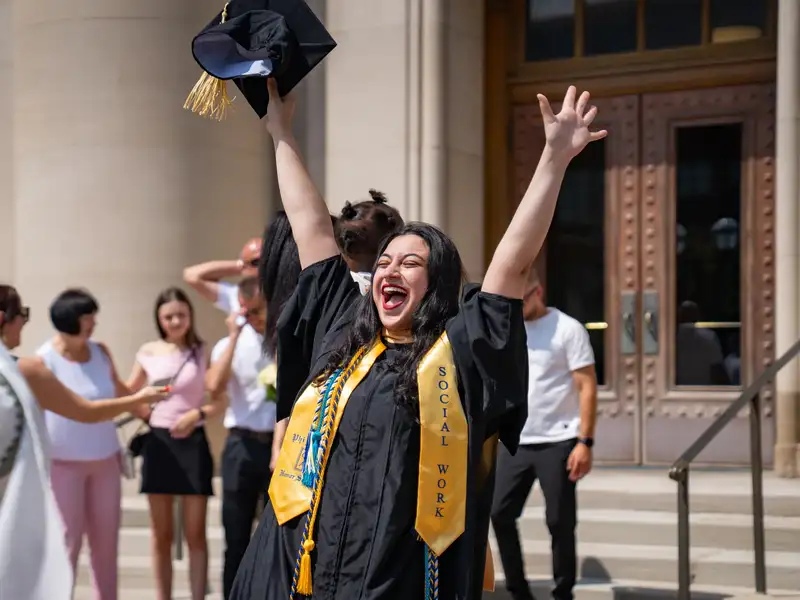 A joyful MSW graduate throws both arms in the air outside a campus building, laughing and holding her cap, wearing a Social Work stole and honor society cords over her black gown.
