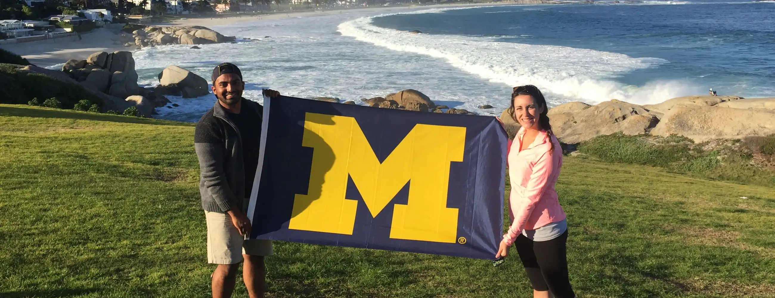 Two students holding the U-M flag in front of a beach.