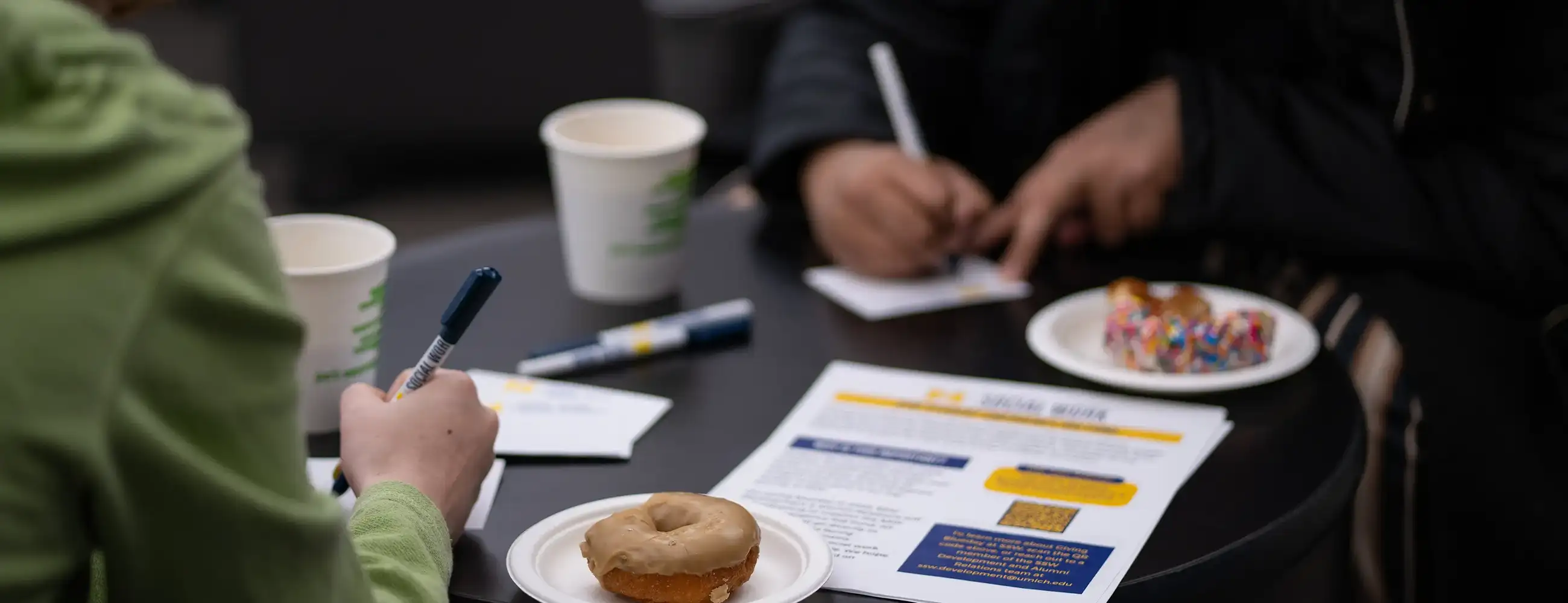 People writing on a table with coffee and donuts.