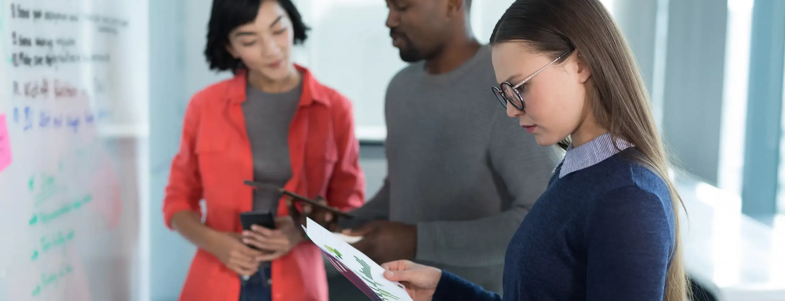 People in an office space looking at a dry erase board with a woman in the foreground reading a report.
