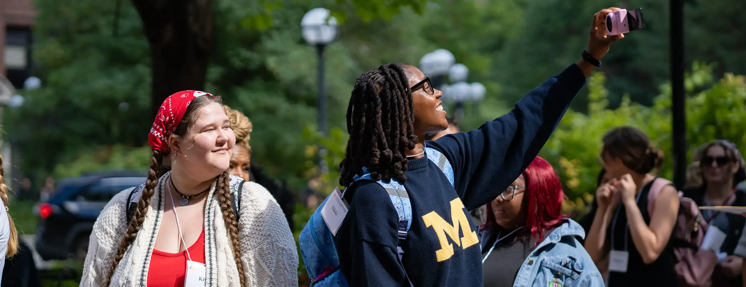 A student wearing a Michigan sweatshirt takes a selfie while standing with other students during an outdoor campus event.