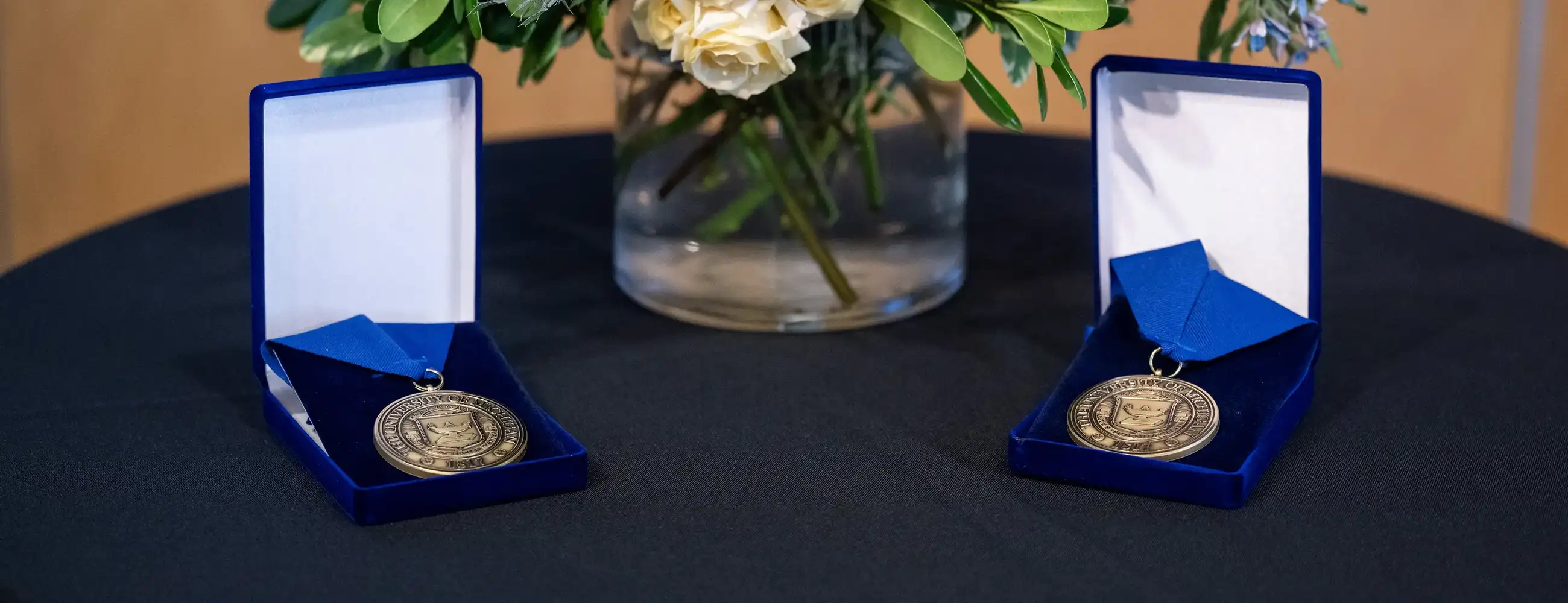 Two ceremonial medals in blue presentation boxes sit on a black table beside a floral arrangement with yellow roses, blue flowers and white roses during the Annmarie Hawkins Professorship installation event.