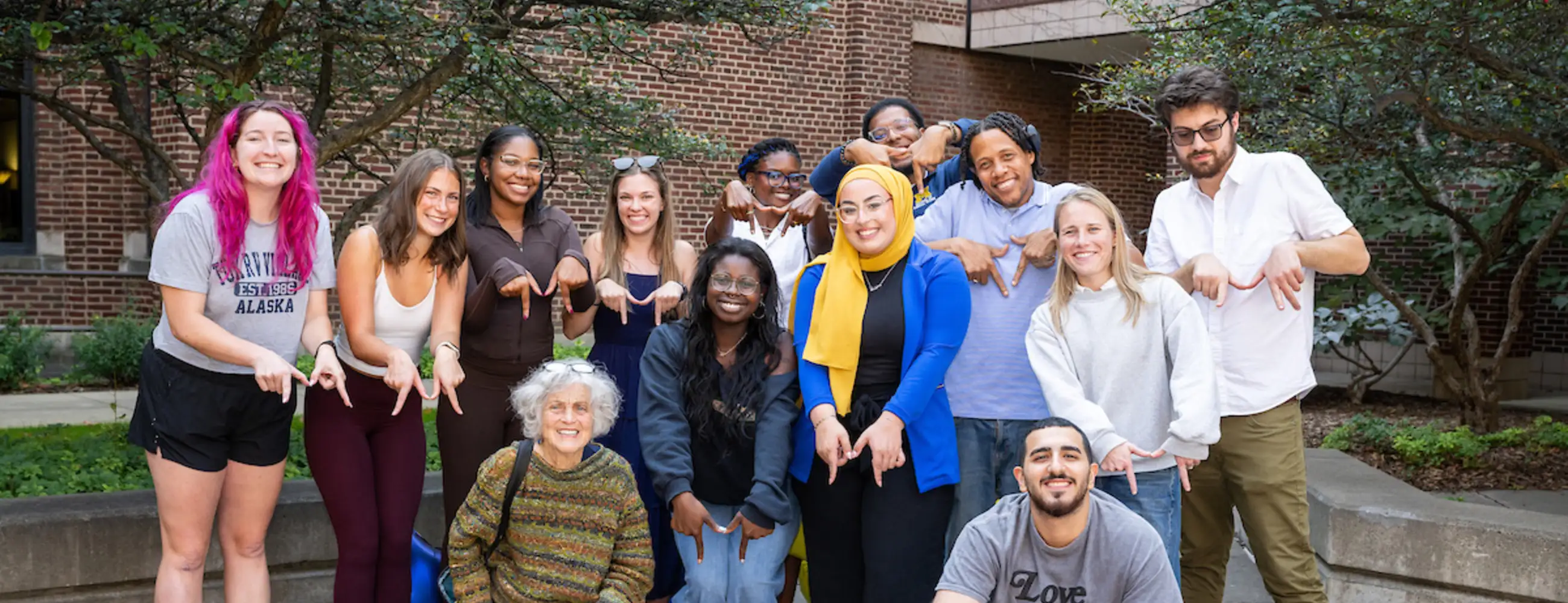 A group of about 14 students, alumni, and faculty pose together in the School of Social Work courtyard, many making the Michigan 'Go Blue' hand sign. Fall leaves dot the brick pavers beneath mature trees.