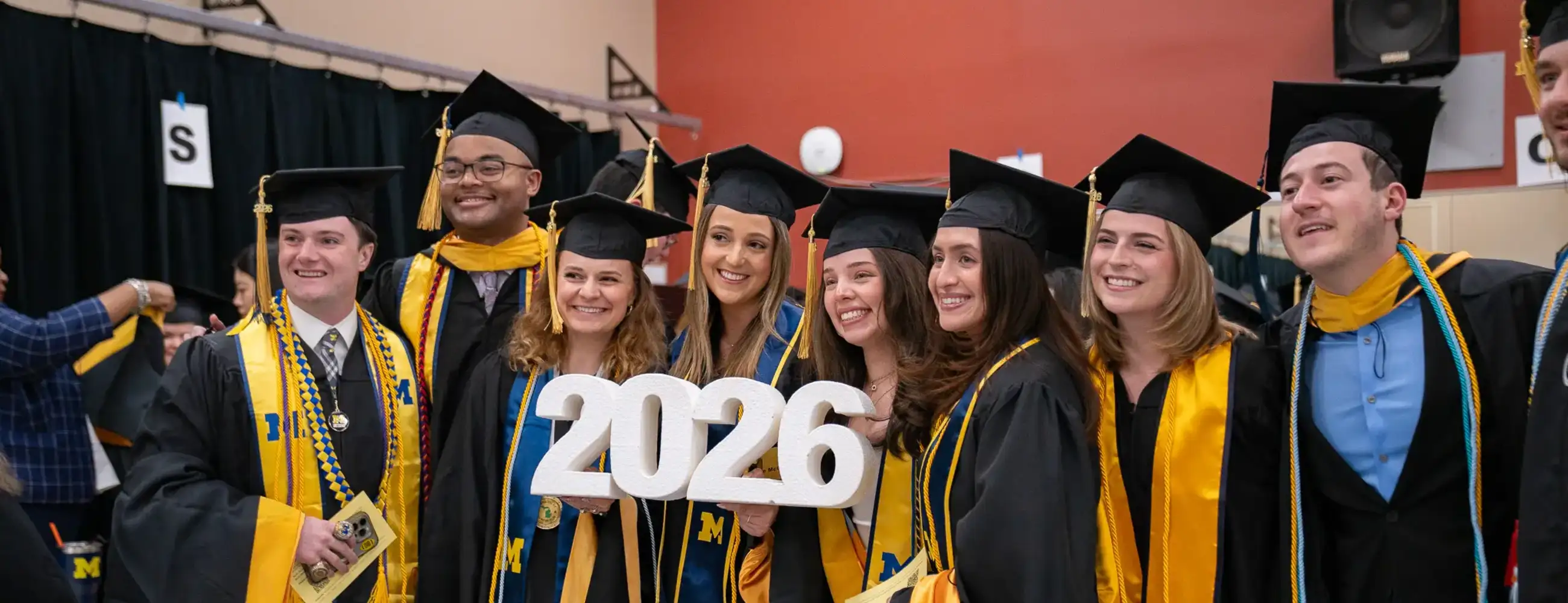 A group of eight University of Michigan School of Social Work graduates in black caps and gowns with maize and blue honor cords and stoles, smiling together for a photo. The two graduates in the center hold a large white "2026" sign.