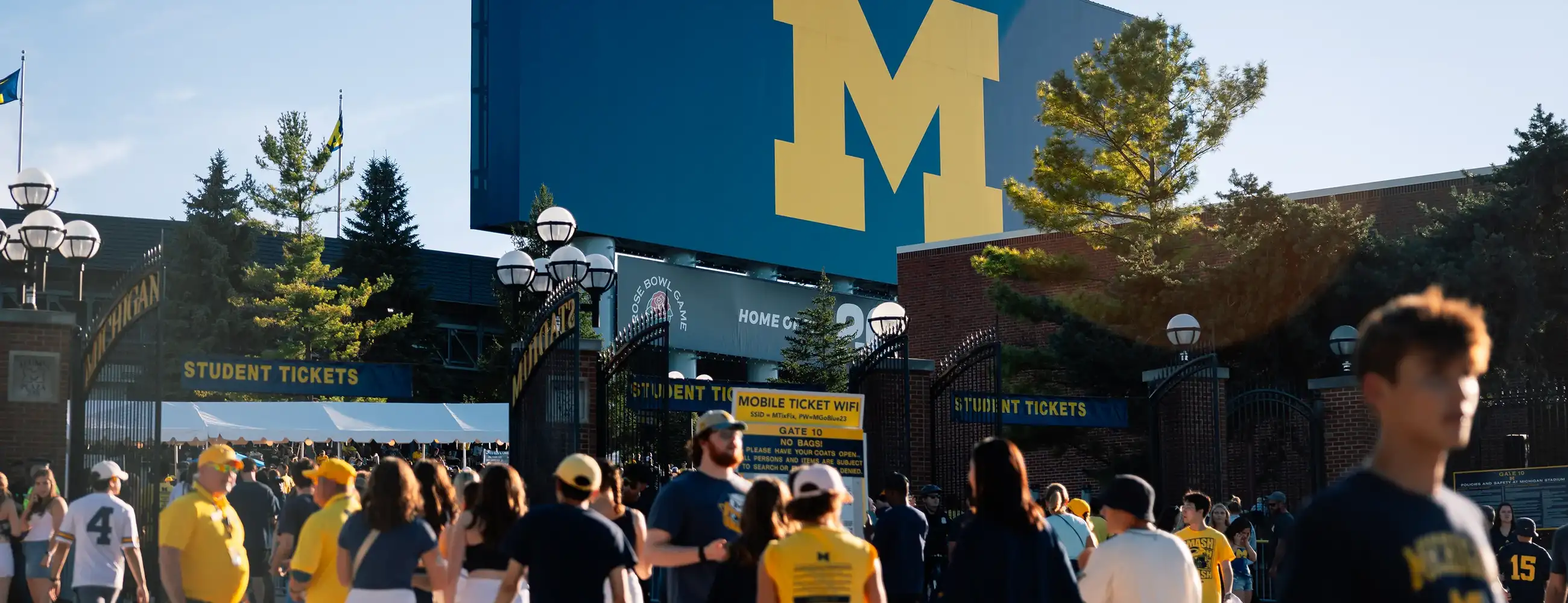 Fans in maize and blue gather outside Michigan Stadium gates on a sunny game day, with a large block M logo visible on the stadium facade.