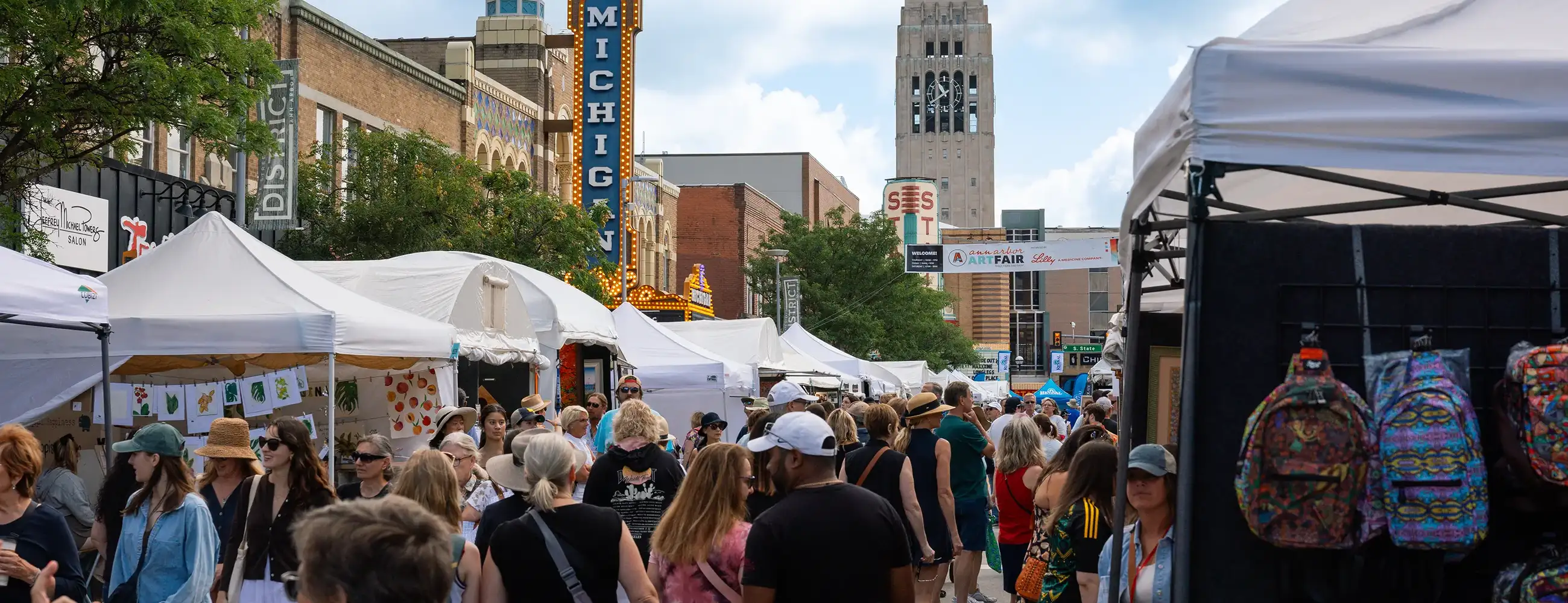 State Street during the Ann Arbor Art Fair, packed with pedestrians browsing white-tented art booths. The Michigan Theater marquee and Burton Memorial Tower are visible in the background under a partly cloudy sky.