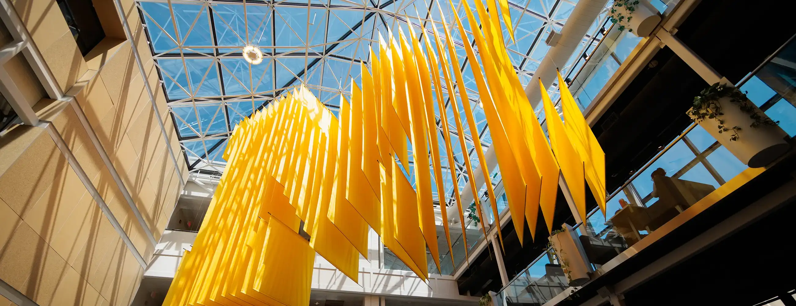 A suspended sculpture of golden-yellow angled panels hangs beneath the glass atrium ceiling of the U-M Student Activities Building.