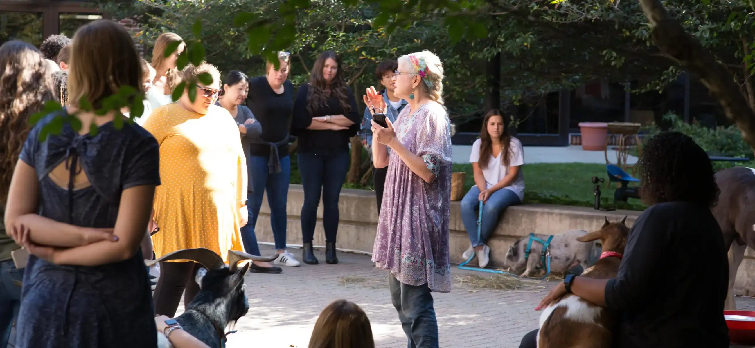 Laura Sanders addresses a group of new students in the University of Michigan School of Social Work courtyard, gesturing as she speaks about her Animal Assisted Interventions course. Several animals, including a goat, a pig, and a small horse, are present among the gathered students.