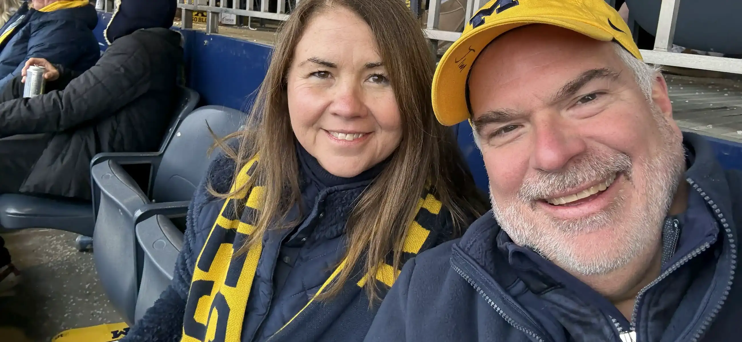 Maureen and Jim Lico smiling at a University of Michigan football game at Michigan Stadium, wearing maize and blue gear including a Michigan "M" cap and a maize and navy scarf.