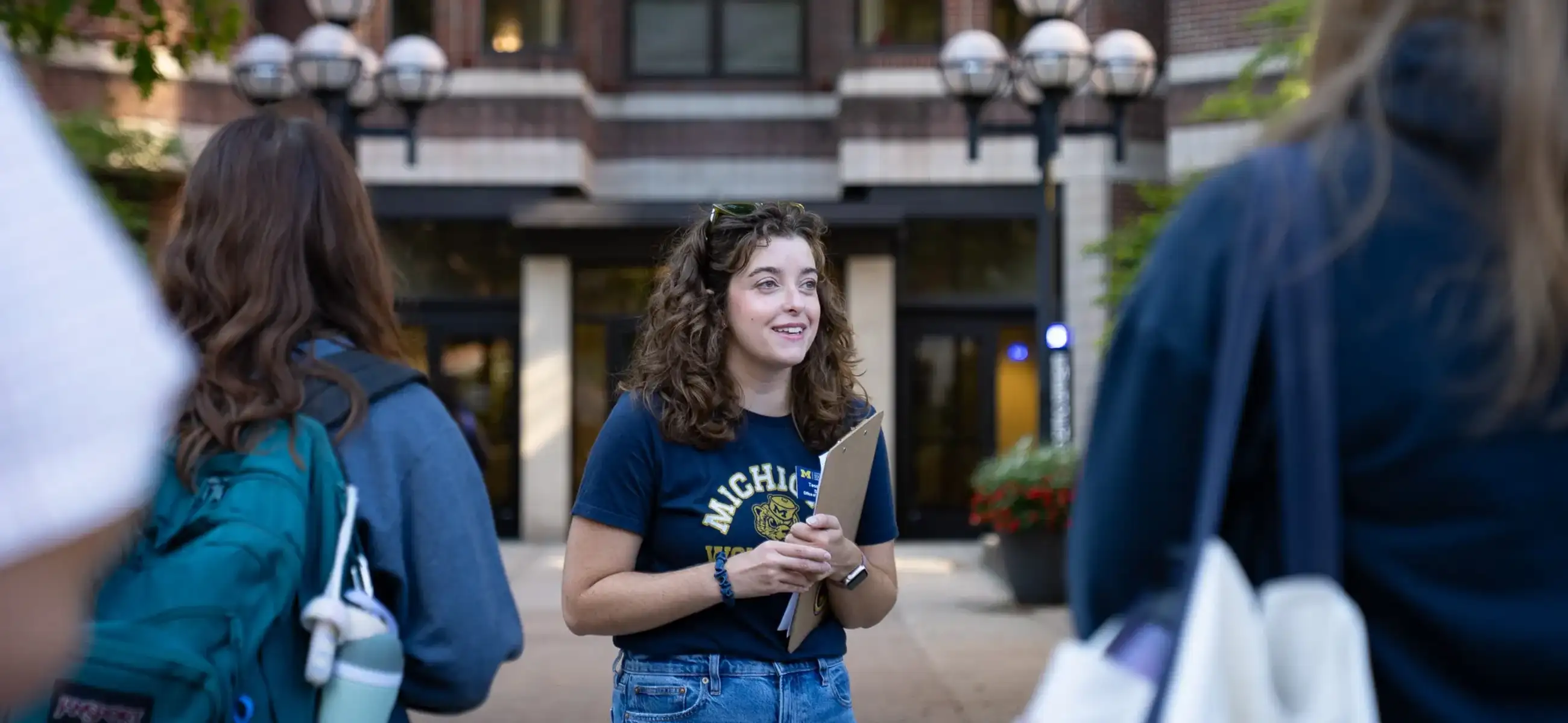 A student tour guide in a navy University of Michigan t-shirt holds a clipboard while speaking to a small group of prospective students outside the School of Social Work building. The brick building's entrance and distinctive globe light fixtures are visible in the background.
