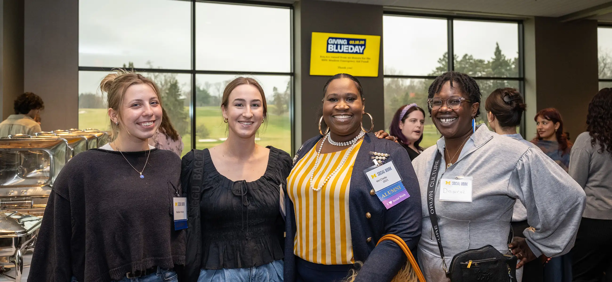Four people pose together smiling at the University of Michigan School of Social Work Month celebration in 2026. They wear name badges, with one displaying an "Alumni" tag. The group stands in a large event space with catering trays and a "Giving Blueday" sign visible in the background, while other attendees mingle behind them.