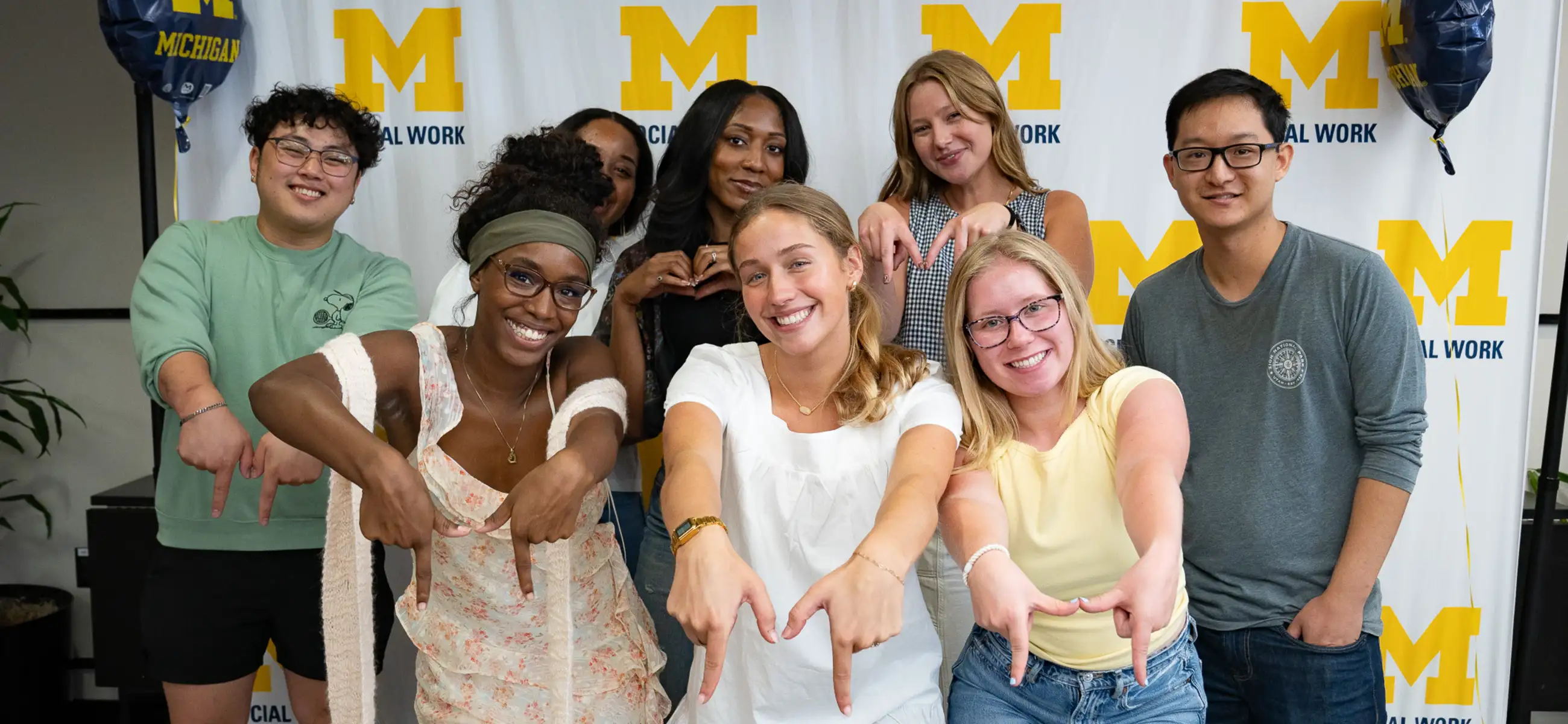 Eight students smiling and forming M hand signs in front of a Michigan Social Work step-and-repeat banner during SEED Week in McGregor Commons.