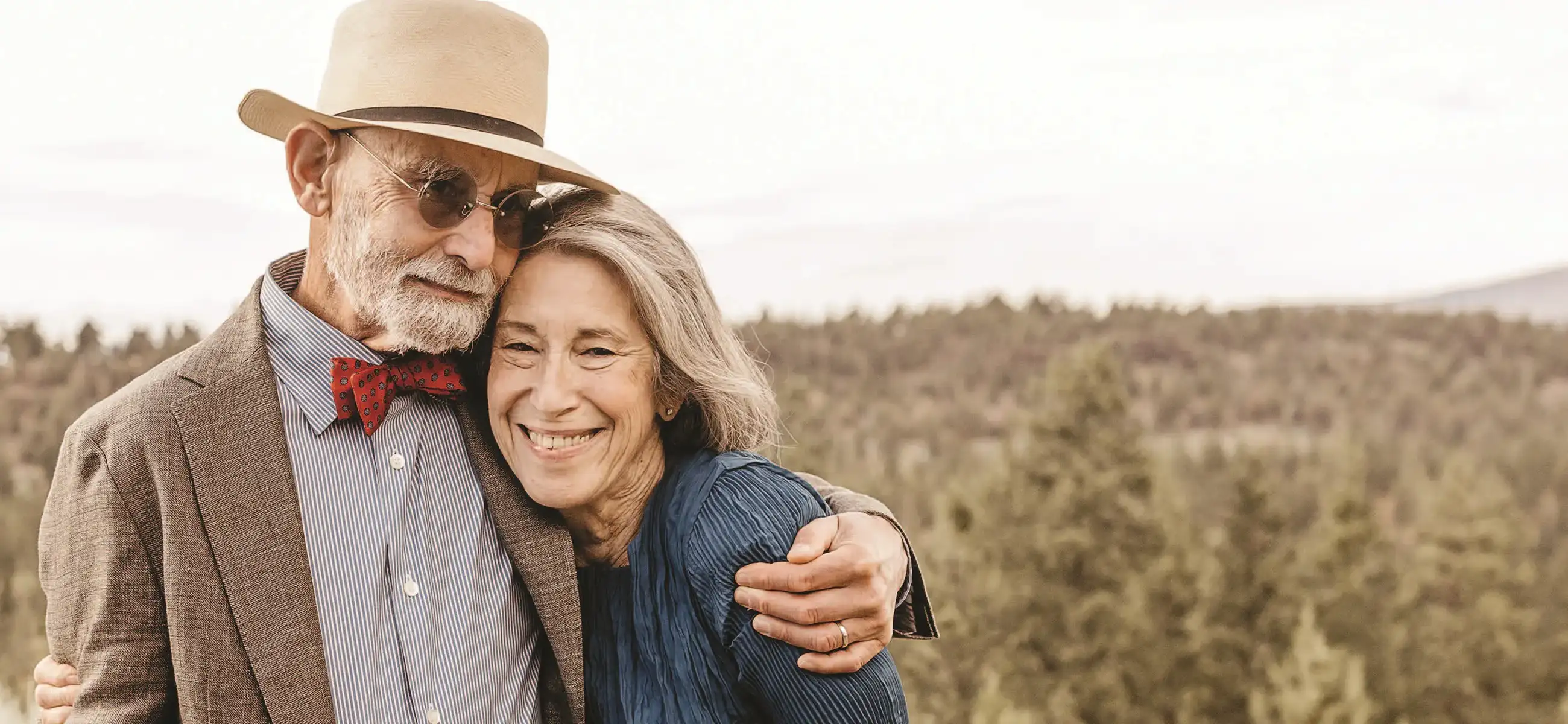 Two people embrace in a sunlit field of dry grass with forested hills behind them.