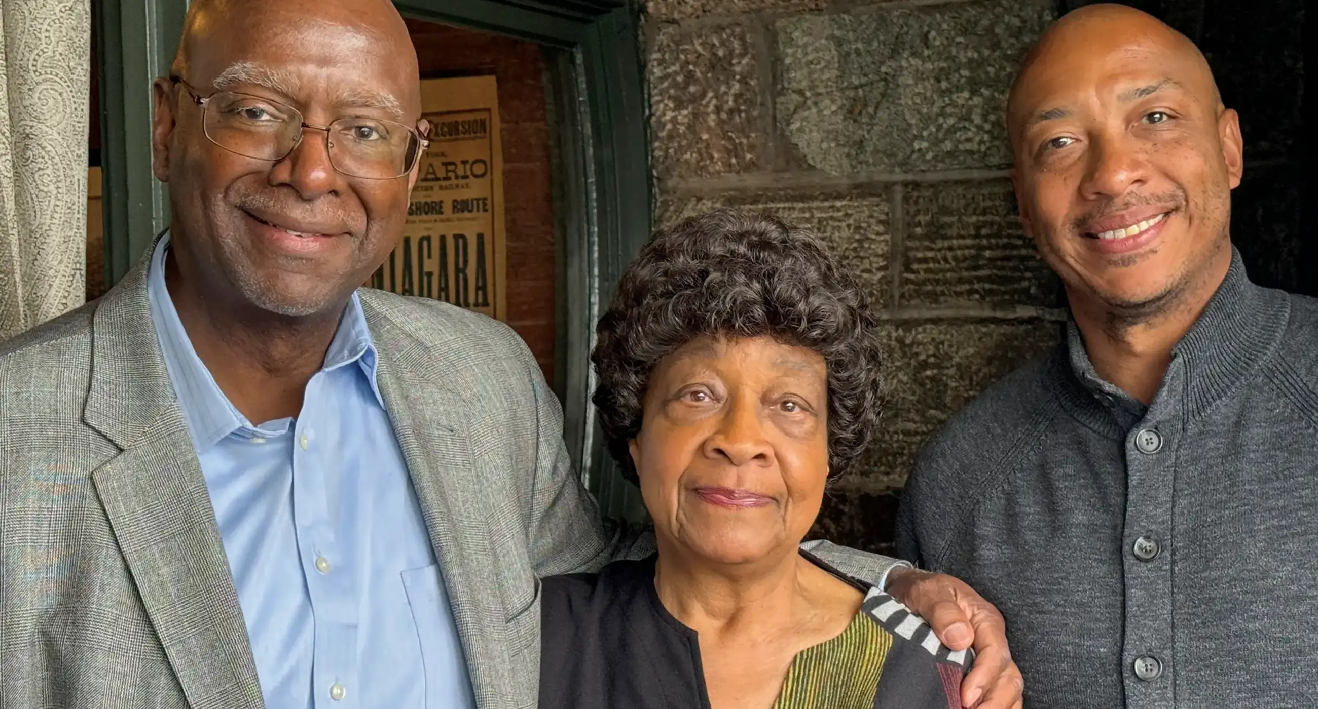 Three people smile for a photo in front of a stone wall and a doorway with a vintage Niagara excursion poster. An older woman in a colorful patterned cardigan stands in the center, with a man in a gray blazer and light blue shirt on the left and a man in a dark gray button-up sweater on the right, his arm around her shoulder.