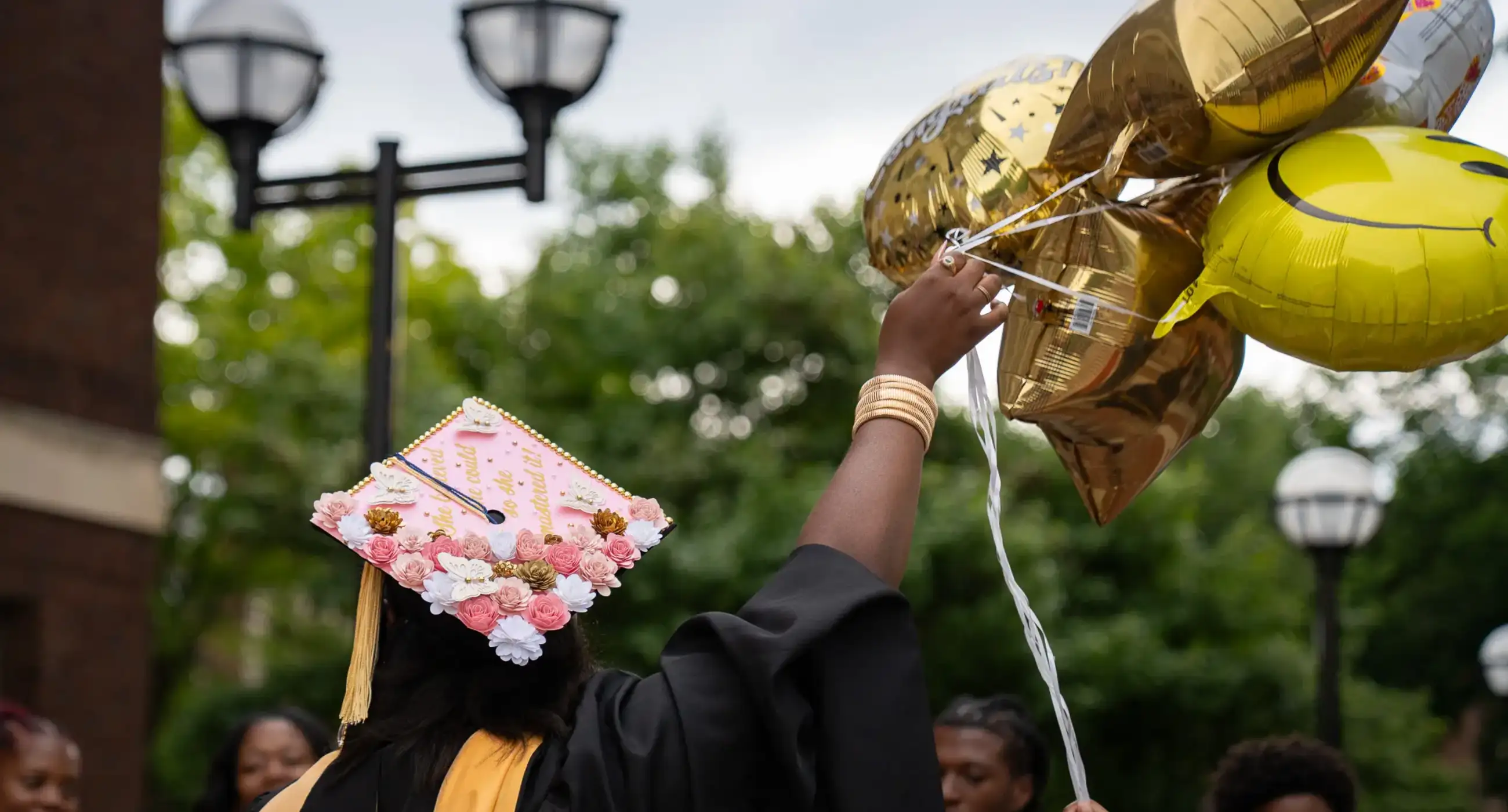 A graduate in a pink cap decorated with paper flowers reaches up to a bunch of gold balloons.