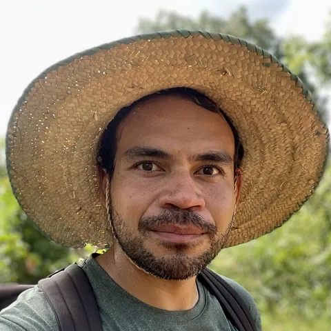 Sevi Fua wearing a large, woven straw hat standing outdoors with green foliage and a partly cloudy sky in the background.