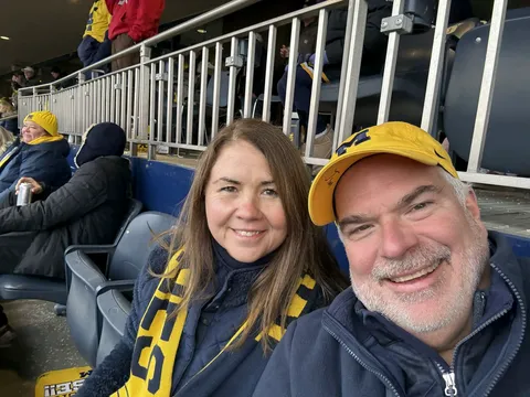 Maureen and Jim Lico smiling at a University of Michigan football game at Michigan Stadium, wearing maize and blue gear including a Michigan "M" cap and a maize and navy scarf.
