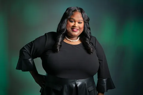 Courtney Smith smiles at the camera with one hand on her hip, wearing a black top with leather-trimmed bell sleeves, a black leather skirt, and a pearl necklace, against a soft green and gray background.