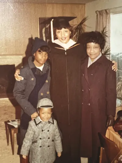 A graduate in cap and gown poses indoors with family members in winter coats, including a young child.