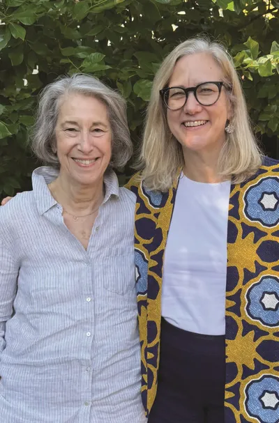 Two women smile together outdoors in front of a green hedge, one with an arm around the other.